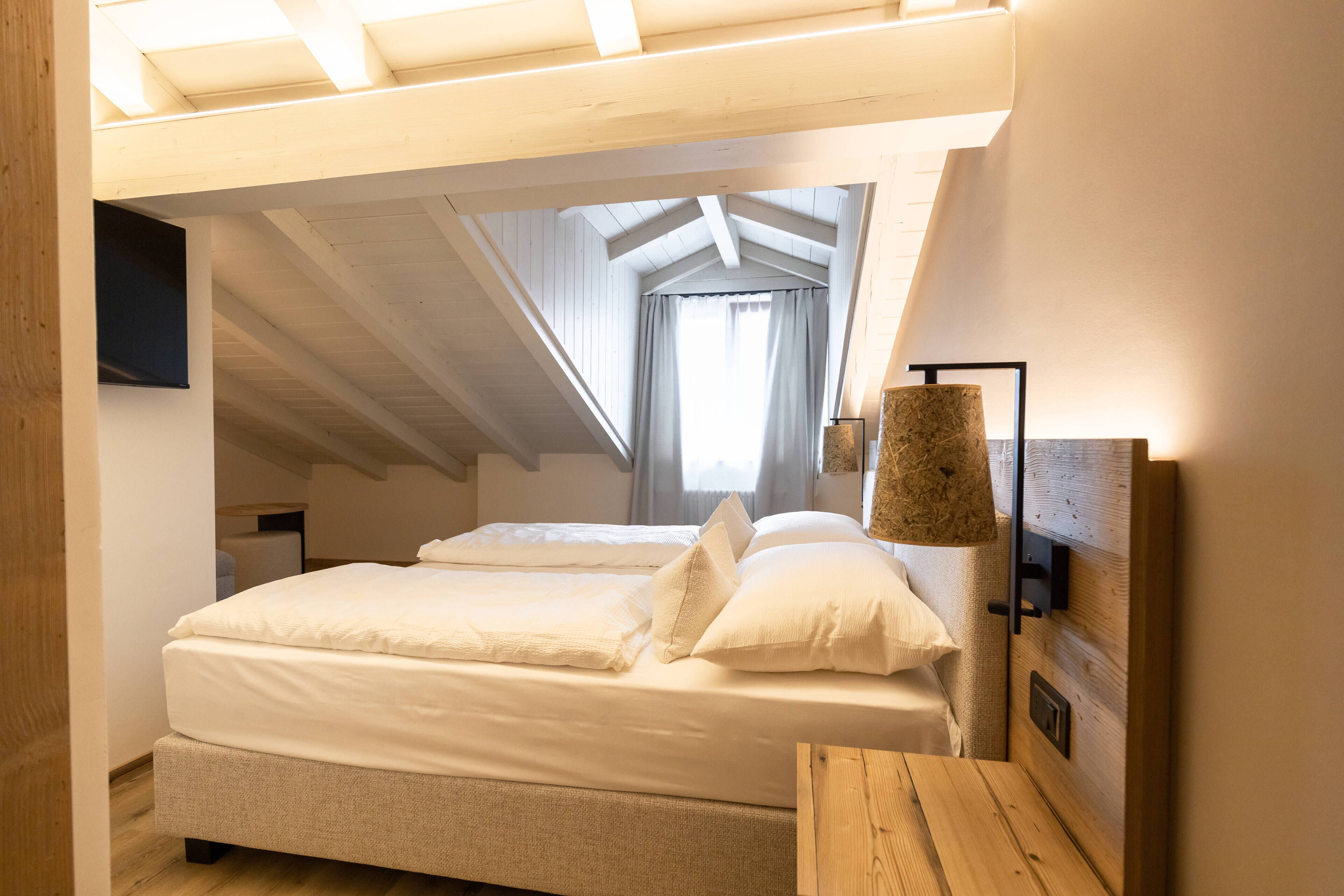 Una camera da letto con soffitto a vista, un letto con biancheria bianca, un comodino in legno e una lampada fissata al muro.