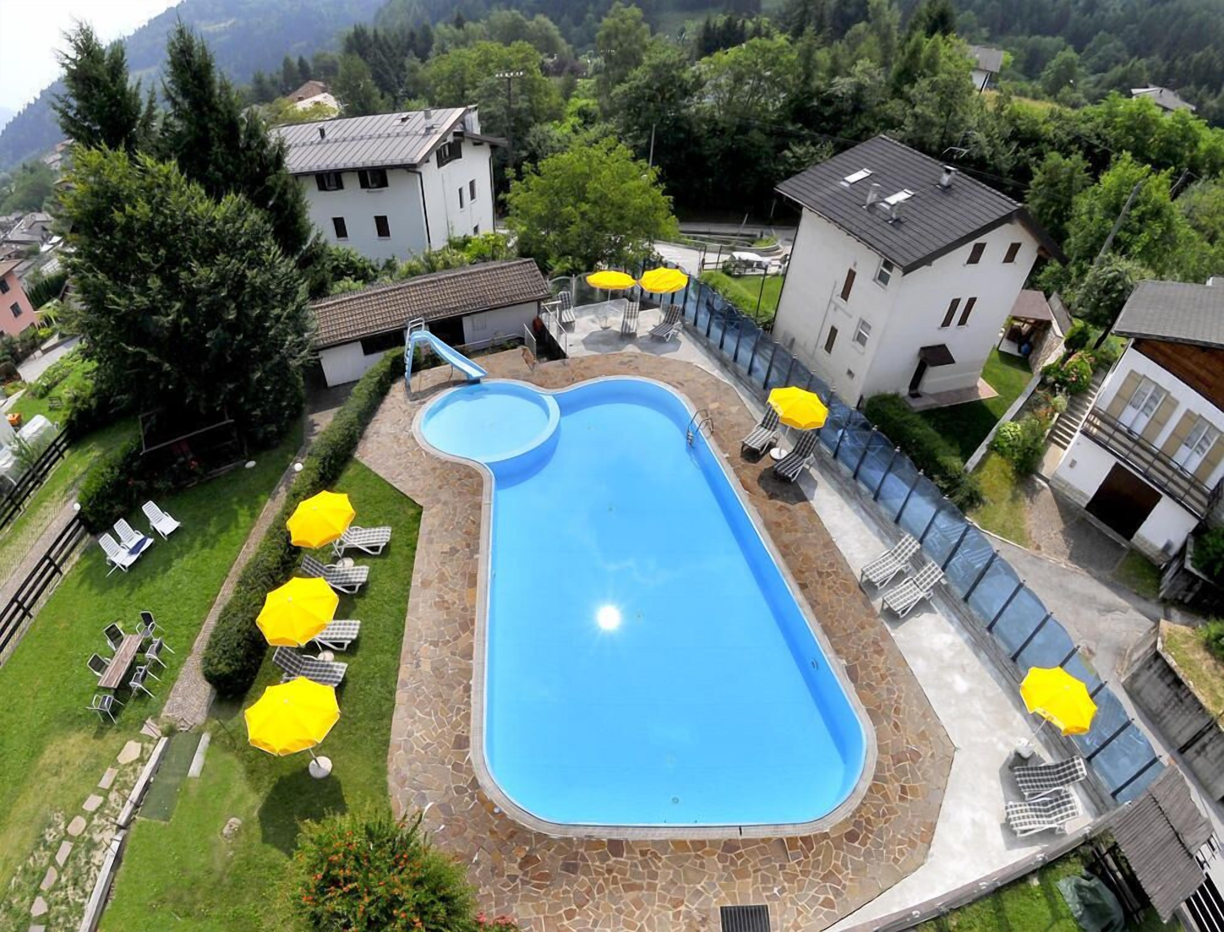 A swimming pool with a slide, surrounded by lounge chairs and umbrellas, set in a residential area with houses and greenery.
