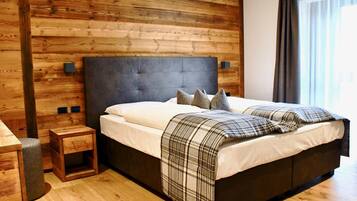 Un dormitorio moderno con cabecera de madera, cama con ropa blanca y manta a cuadros, una mesita de noche de madera y una ventana con cortinas.