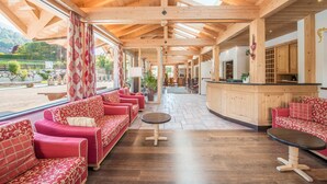 A spacious lobby with wooden beams, red patterned sofas, a central round table, and a kitchen area with wooden cabinets. - Hotel Alpenrose (Altopiano della Vigolana)