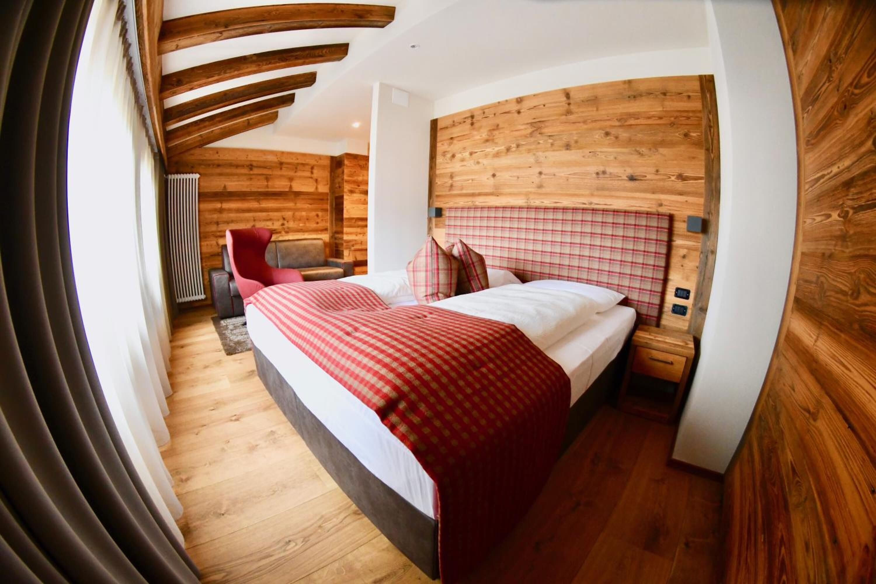 A modern hotel room with a wooden headboard, a large bed with a red and white checkered bedspread, a wooden floor, and a seating area with a chair and a small table.