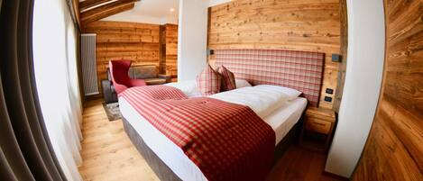 A modern hotel room with a wooden headboard, a large bed with a red and white checkered bedspread, a wooden floor, and a seating area with a chair and a small table.