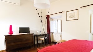 A bedroom with a bed, a desk with a computer, a chair, a red vase, and a window with curtains. - Spanish Steps Studio Apartment (Rome)