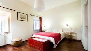 A bedroom with a bed, a bedside table, a lamp, a red chest of drawers, a wicker basket, and a framed picture on the wall. - Spanish Steps Studio Apartment (Rome)