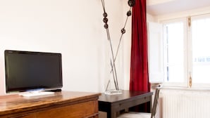 A room with a wooden desk, a television on a stand, a chair, a radiator, and a window with a red curtain. - Spanish Steps Studio Apartment (Rome)