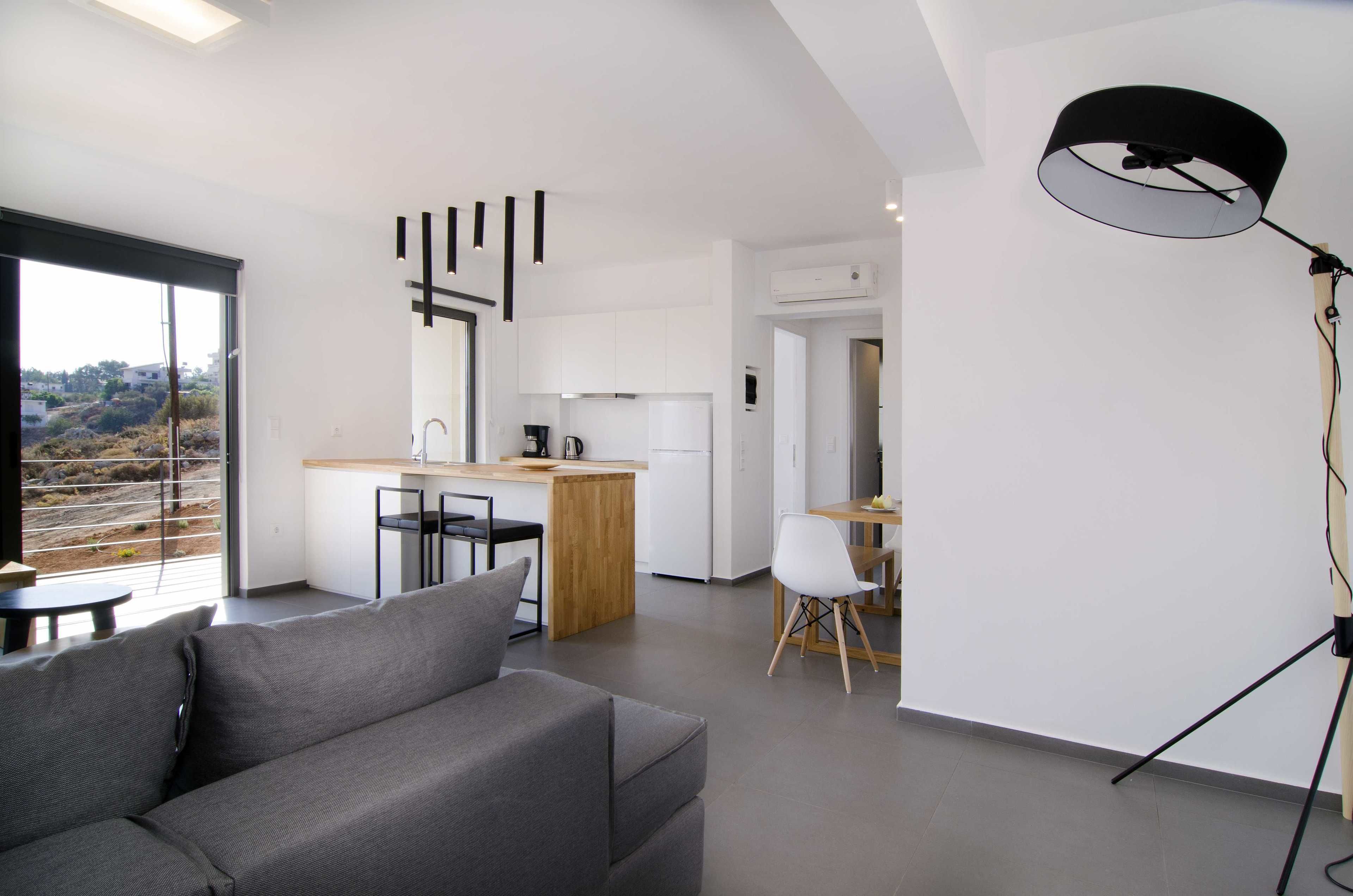A modern interior with a grey sofa, a dining area, a kitchen, and a large window offering a view of the outdoors.