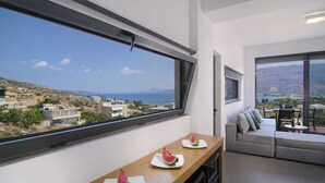 Une chambre moderne dotée d’une grande fenêtre donnant sur un paysage côtier, une table en bois avec des tranches de pastèque, et un canapé gris offrant une vue agréable.