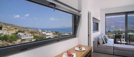 Une chambre moderne dotée d’une grande fenêtre donnant sur un paysage côtier, une table en bois avec des tranches de pastèque, et un canapé gris offrant une vue agréable.