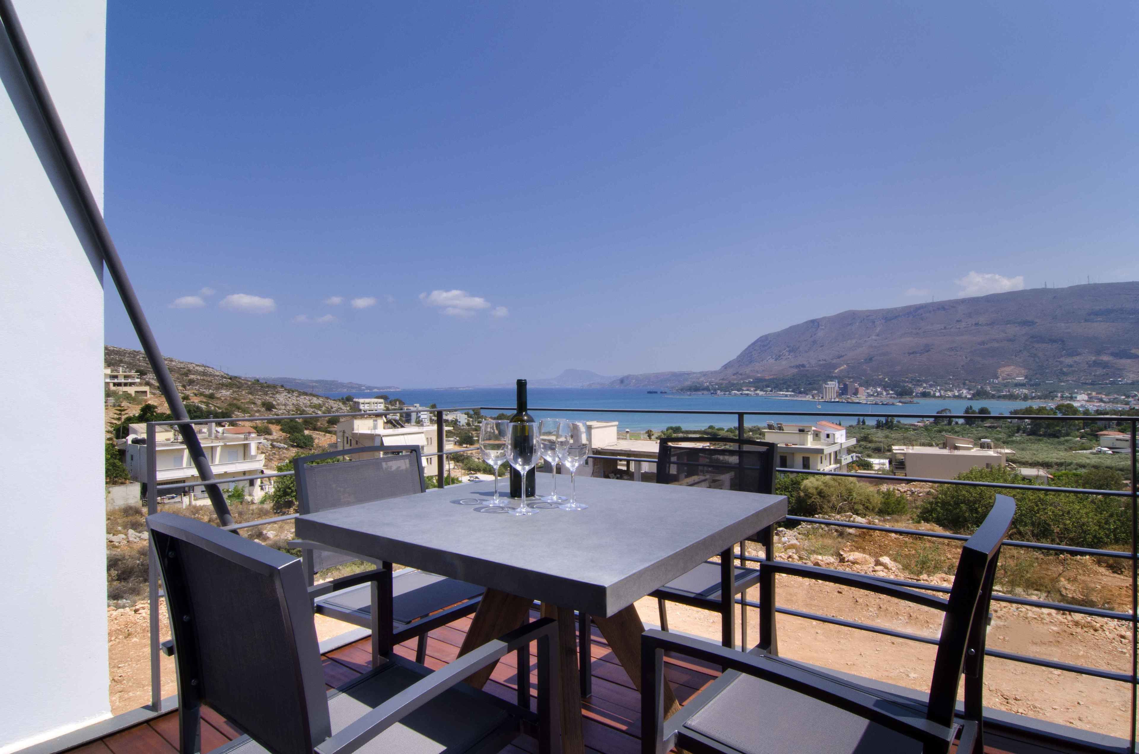 Une table entourée de chaises sur un balcon donnant sur une vue côtière, avec des bâtiments et des collines visibles au loin.