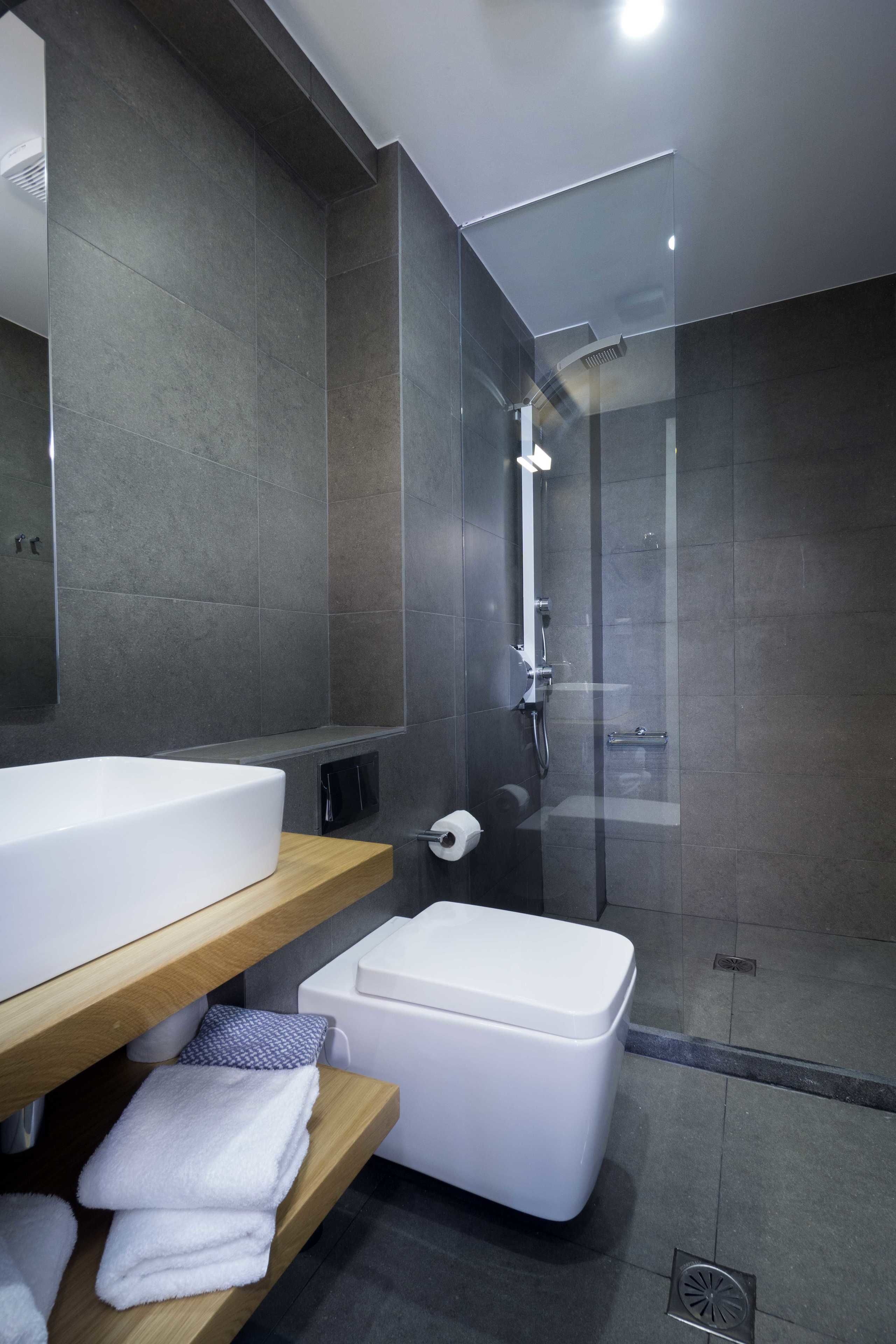 Une salle de bain moderne avec un grand miroir, un lavabo blanc, une douche en verre et une étagère en bois où sont posées des serviettes.