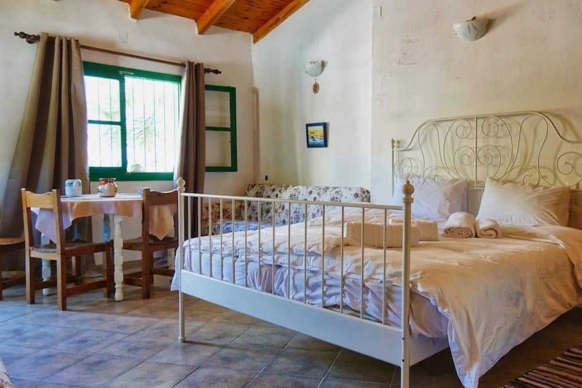 A bedroom with a metal bed frame, a wooden chair, a table with a pitcher, and a green window.