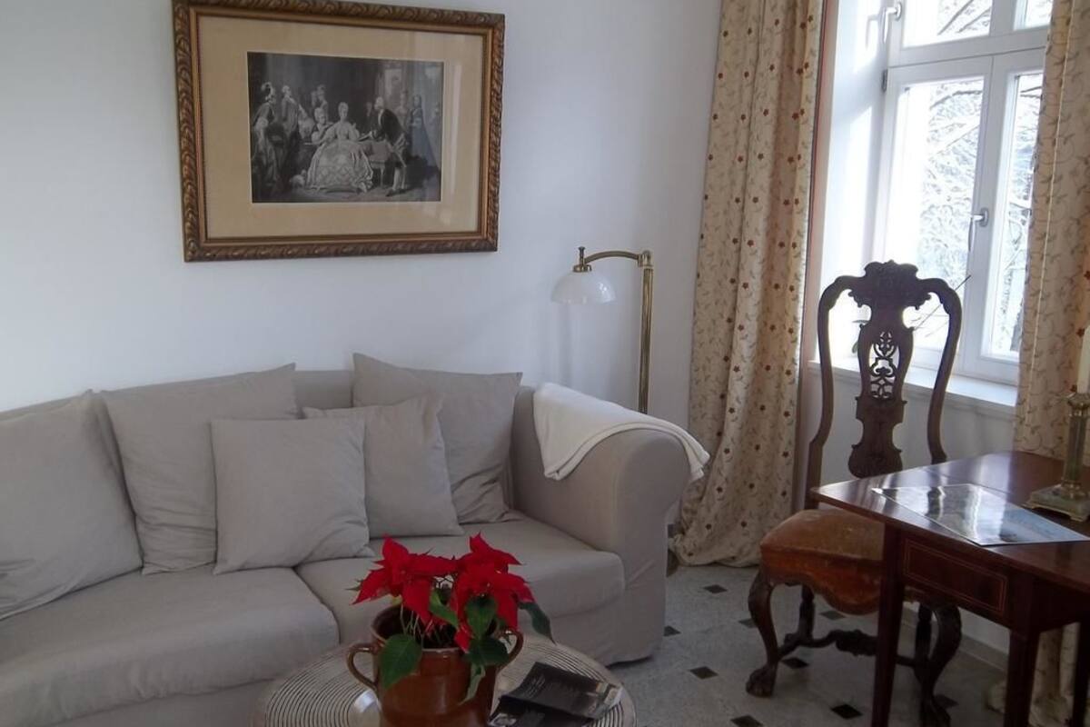 Una sala de estar con un sofá, una mesa de centro con una planta en maceta, un cuadro enmarcado en la pared, una lámpara de pie y una ventana con cortinas.