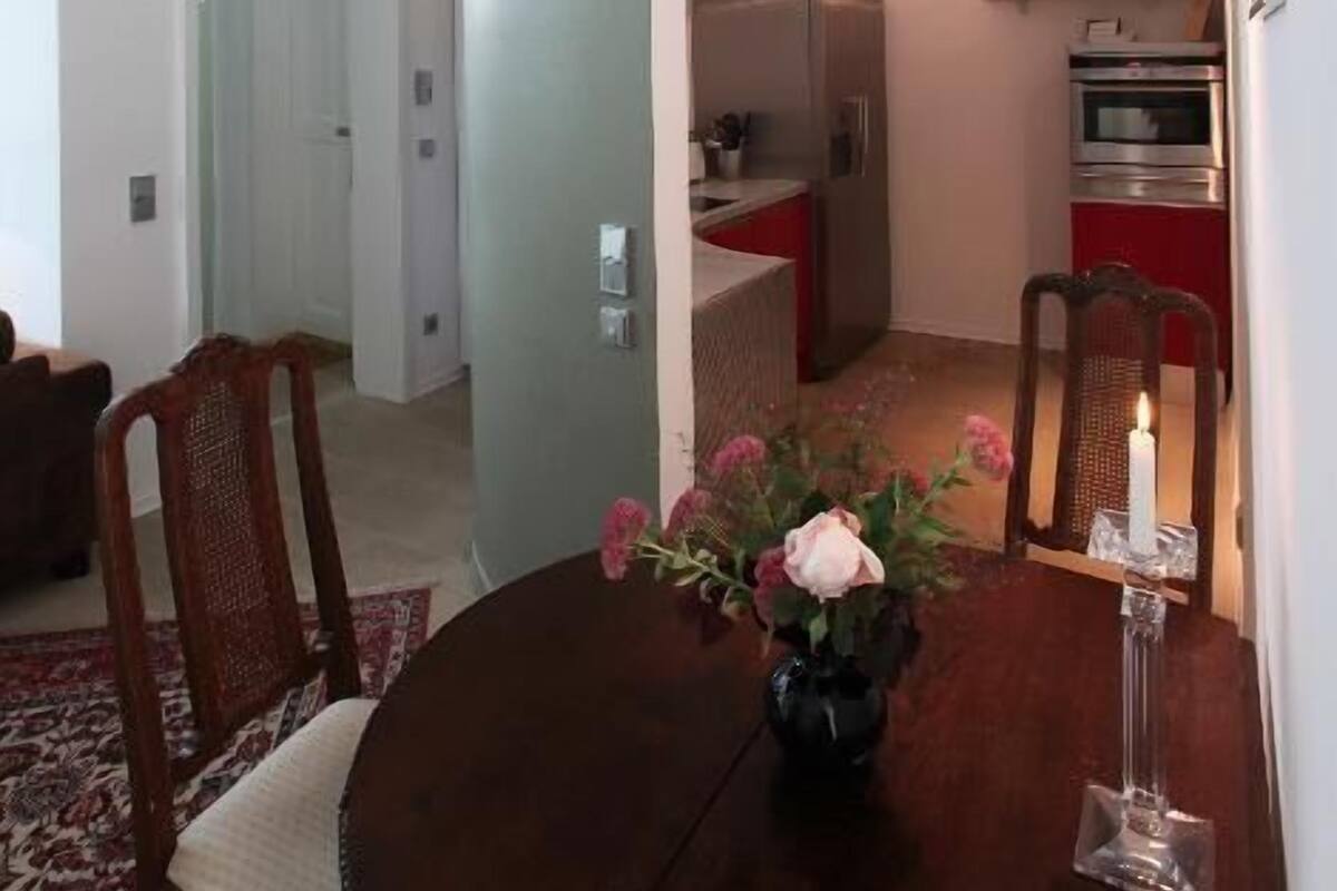Un espacio habitable compacto con comedor que incluye una mesa redonda, un florero con flores y una vela. Se ve una cocina al fondo con electrodomésticos modernos.