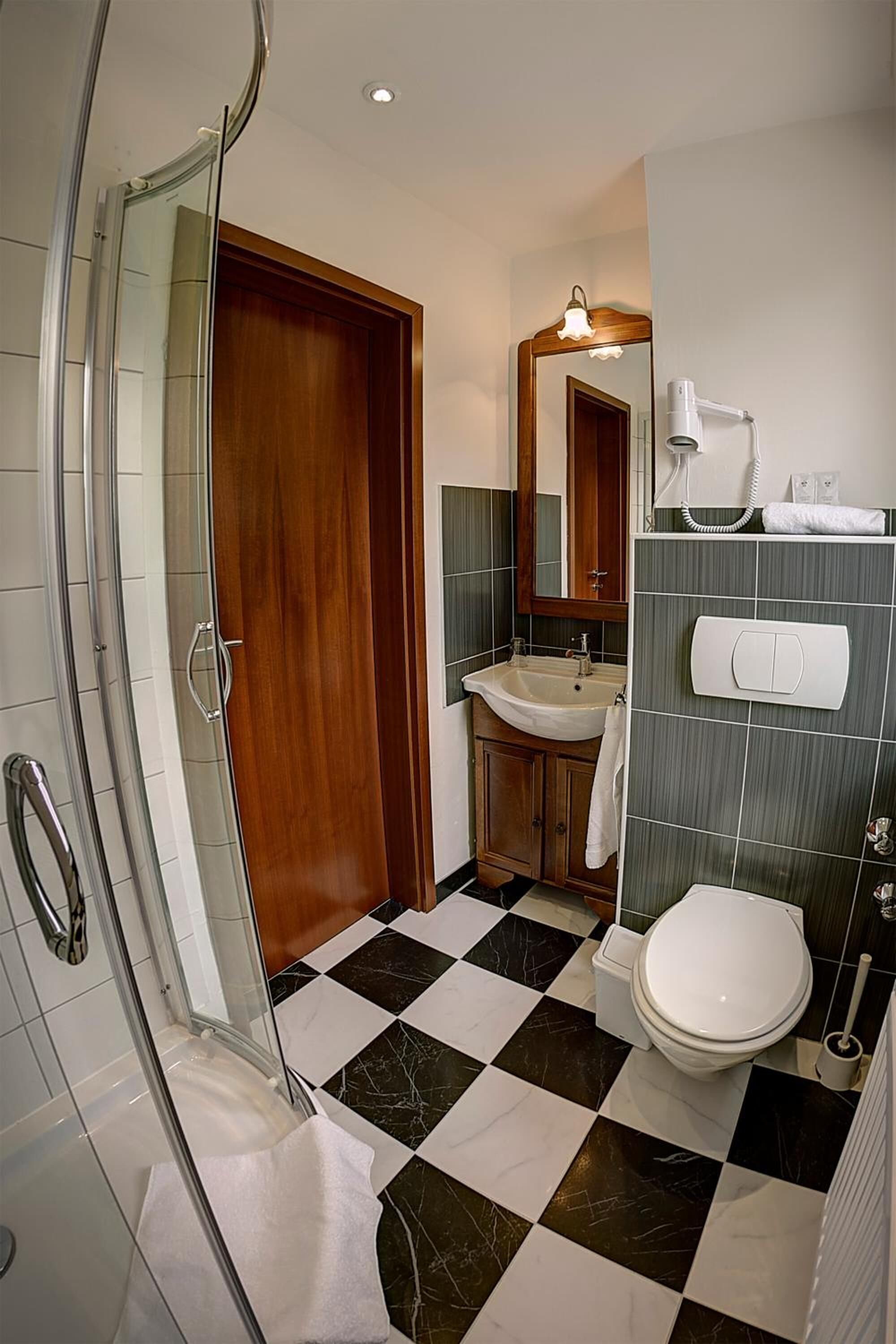 A bathroom with a glass shower enclosure, a wooden door, a sink with a mirror above it, a toilet, and a tiled floor.