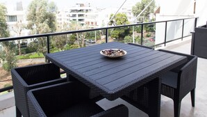 En balkon med et mørkt fletbord og stole, en skål med frugt og udsigt til bygninger og træer.
