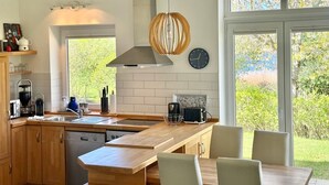 A modern kitchen with a central island, a built-in dishwasher, and a large window offering a view of greenery. - Quiet 5* vacation home right on the water | WLAN | sauna | fireplace | whirlpool tub (Kosel / Schlei)