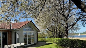 A house with a patio, a table, and chairs, near a tree and a body of water. - Quiet 5* vacation home right on the water | WLAN | sauna | fireplace | whirlpool tub (Kosel / Schlei)