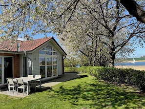 A house with a patio, a table, and chairs, near a tree and a body of water.