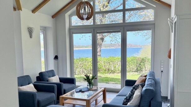 A modern living room with a large window, a wooden ceiling, and a view of the sea. - Quiet 5* vacation home right on the water | WLAN | sauna | fireplace | whirlpool tub (Kosel / Schlei)