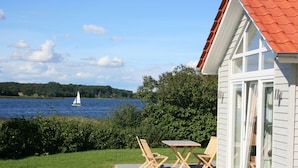 Een huis met een terras dat uitkijkt over een meer, met een tafel en stoelen, en een zeilboot in de verte.