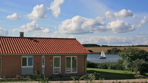 Een bakstenen huis met een rode pannendak, een kleine tuin en een meer met een zeilboot op de achtergrond.
