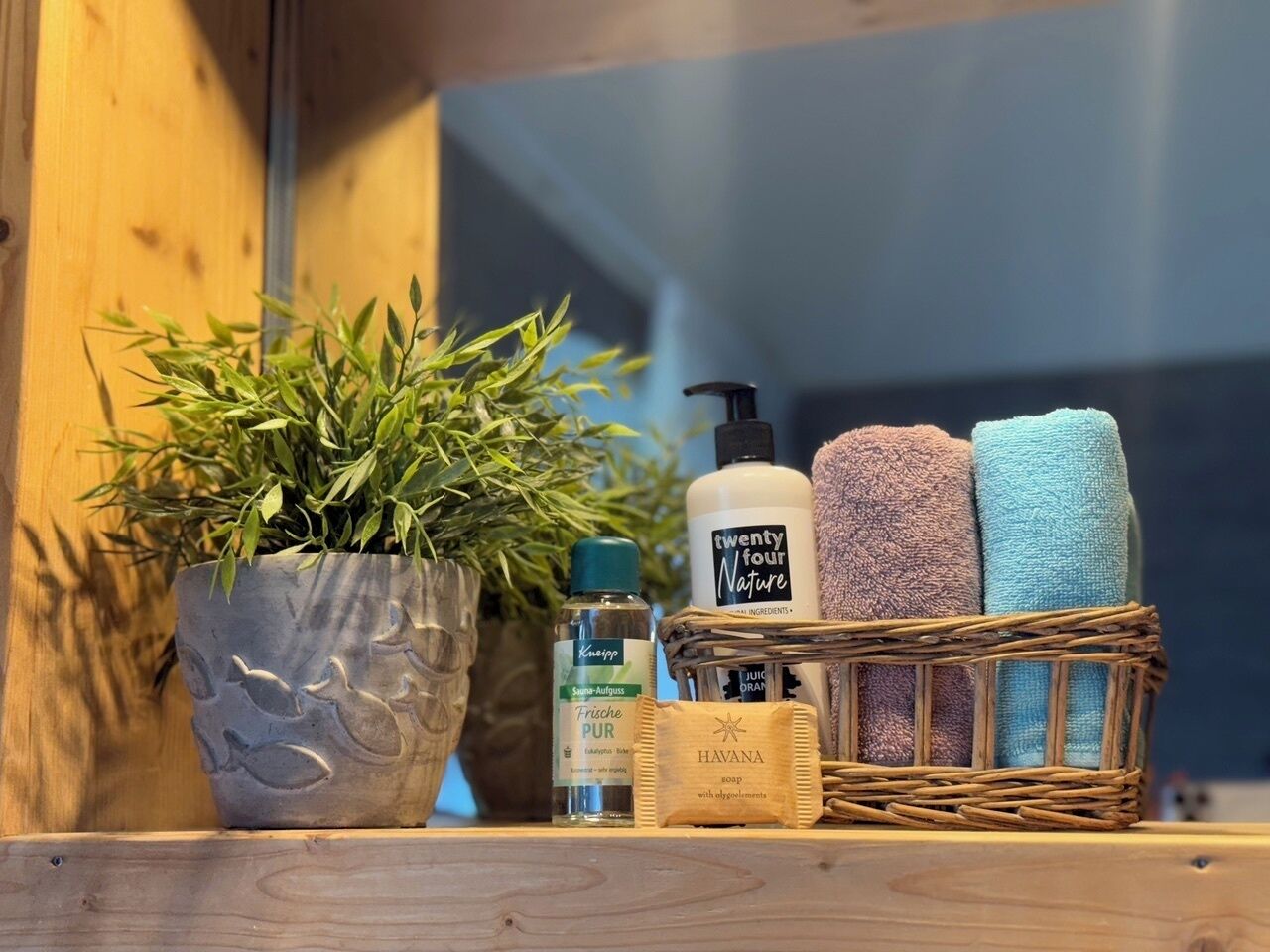 Ein Holzregal mit einem Blumentopf, einer Flasche Frische Aufguss, einer Flasche Havana Seife und einem Korb mit Handtüchern.