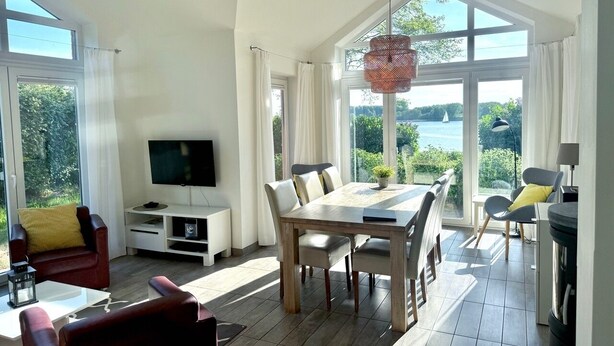 A spacious living room with a large dining table, comfortable seating, a flat-screen TV, and a view of greenery and water through large windows. - Holiday house in direct water situation | Sauna | Whirlpool | Fireplace | Free Wi-Fi (Kosel)