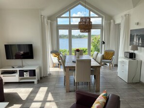 A modern living room with a large window overlooking a body of water, a dining area with a table and chairs, a television on a stand, and a sofa. - Schlei Resort Marina sleeves (Kosel)