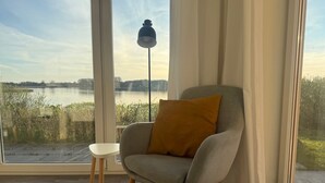 Une pièce avec vue sur un lac, une chaise avec un coussin, une petite table et un lampadaire.