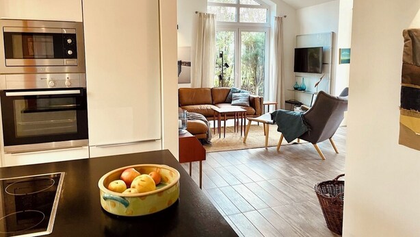 A modern kitchen with a bowl of fruit on the counter, a dining area with a sofa and chairs, and a TV in the background. - Schlei Resort Marina sleeves (Kosel)