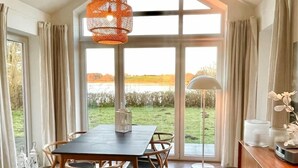 A dining area with a table and chairs, a hanging orange light fixture, and a view of a field through large windows. - Schlei Resort Marina sleeves (Kosel)