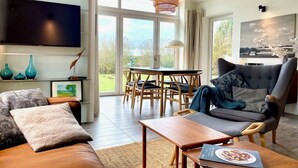 A modern living room with a sofa, armchair, dining table, and a flat-screen TV. - Schlei Resort Marina sleeves (Kosel)