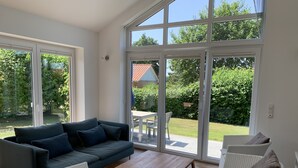 Ein geräumiges Wohnzimmer mit einem dunkelblauen Sofa, weißen Korbstühlen und einem Holztisch. Große Fenster bieten Blick auf einen Garten und ein Gebäude mit rotem Dach.