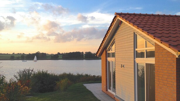 Una casa con techo de tejas, ventanas amplias y un sendero que lleva a un lago con un velero.