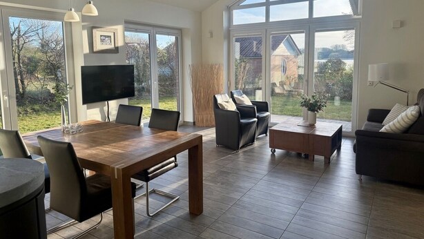 Un salon spacieux avec une table à manger en bois, des chaises en cuir noir, un canapé et un téléviseur. De grandes fenêtres offrent une vue sur l’extérieur.