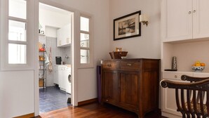 Una habitación con piso de madera, un armario de madera, una silla blanca y una cocina visible a través de una puerta abierta.