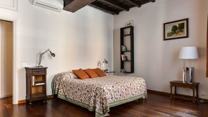 Un dormitorio con piso de madera, una cama con colcha estampada, un mesita de noche con lámpara, una estantería y un cuadro en la pared.