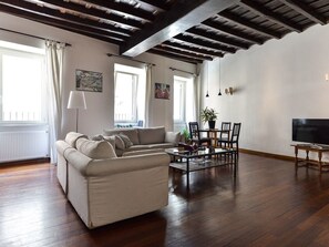 A living room with a sofa, a coffee table, and a TV. There are windows with curtains, a radiator, and a plant. - Spanish Steps Apartment (Rome)