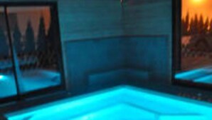 Une piscine intérieure moderne avec des parois en verre transparent, un élément décoratif aquatique illuminé de lumière bleue et une vue sur une forêt à travers le verre.