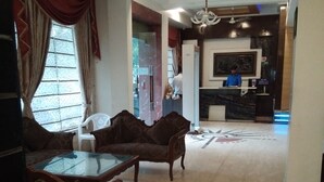 A spacious living area with a sofa set, a glass coffee table, and a patterned armchair. There is a reception desk in the background with a person standing behind it. - Hotel Blessings (New Delhi)