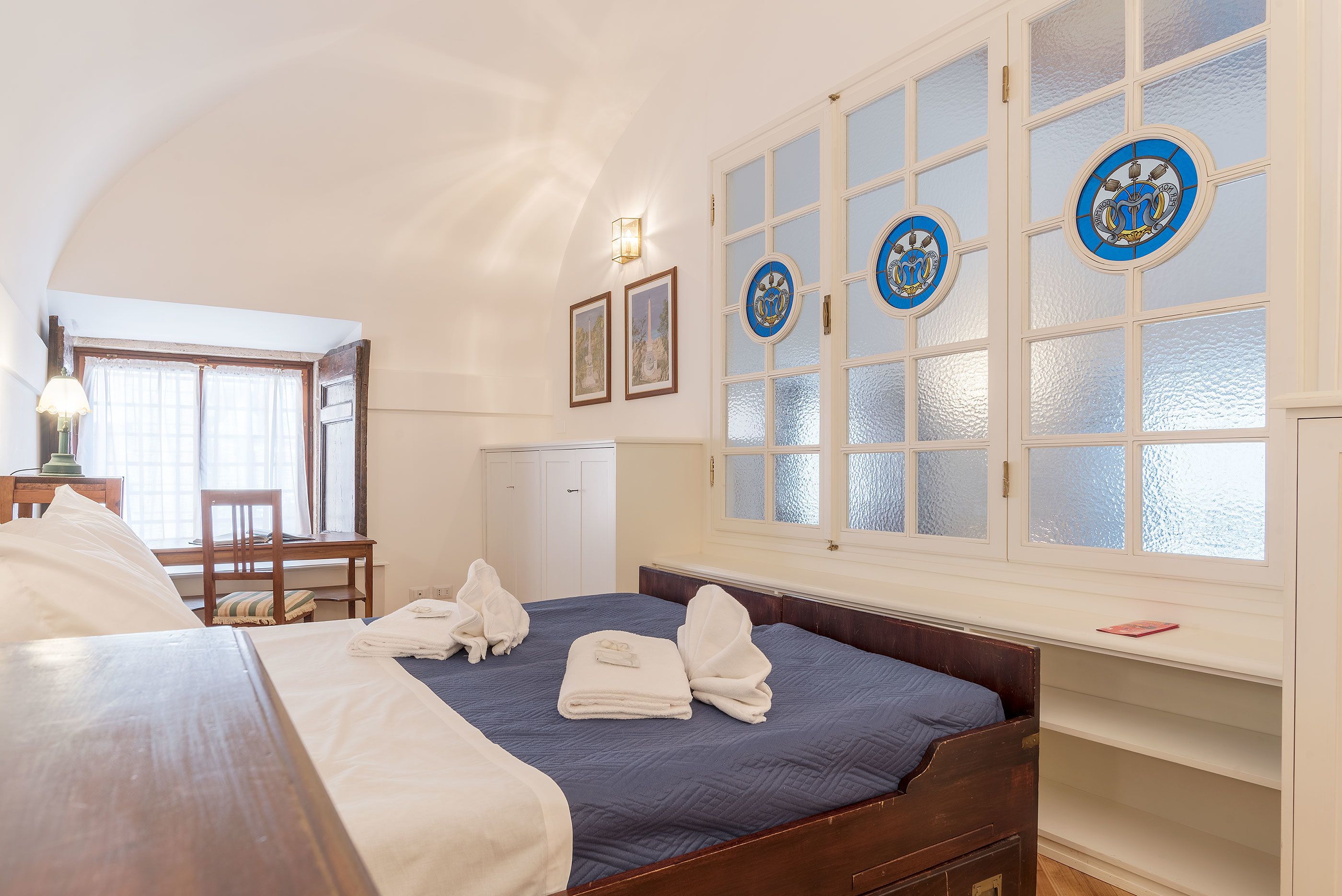 Ein Hotelzimmer mit Bett, Schreibtisch, Stuhl und einem Fenster mit dekorativen Glasscheiben.
