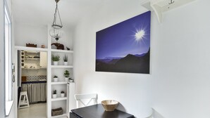 Ein Essbereich mit einem schwarzen Tisch und weißen Stühlen, einer Holzschüssel auf dem Tisch sowie einem an der Wand hängenden Gemälde einer Berglandschaft.