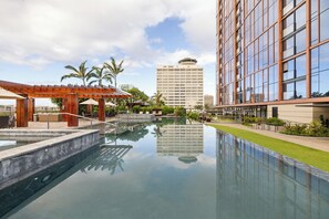 A swimming pool with a wooden pergola and lounge chairs, surrounded by greenery and a tall building in the background.