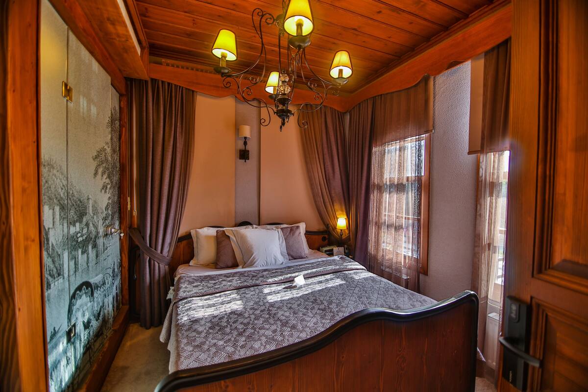A bedroom with a wooden ceiling, a bed with a patterned bedspread, a nightstand with a lamp, and a wall-mounted light.
