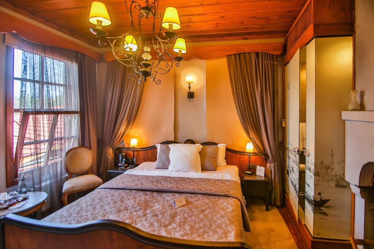 A hotel room with a wooden ceiling, a large bed with a patterned bedspread, two bedside tables with lamps, a chair, and a view of a cityscape through a window.