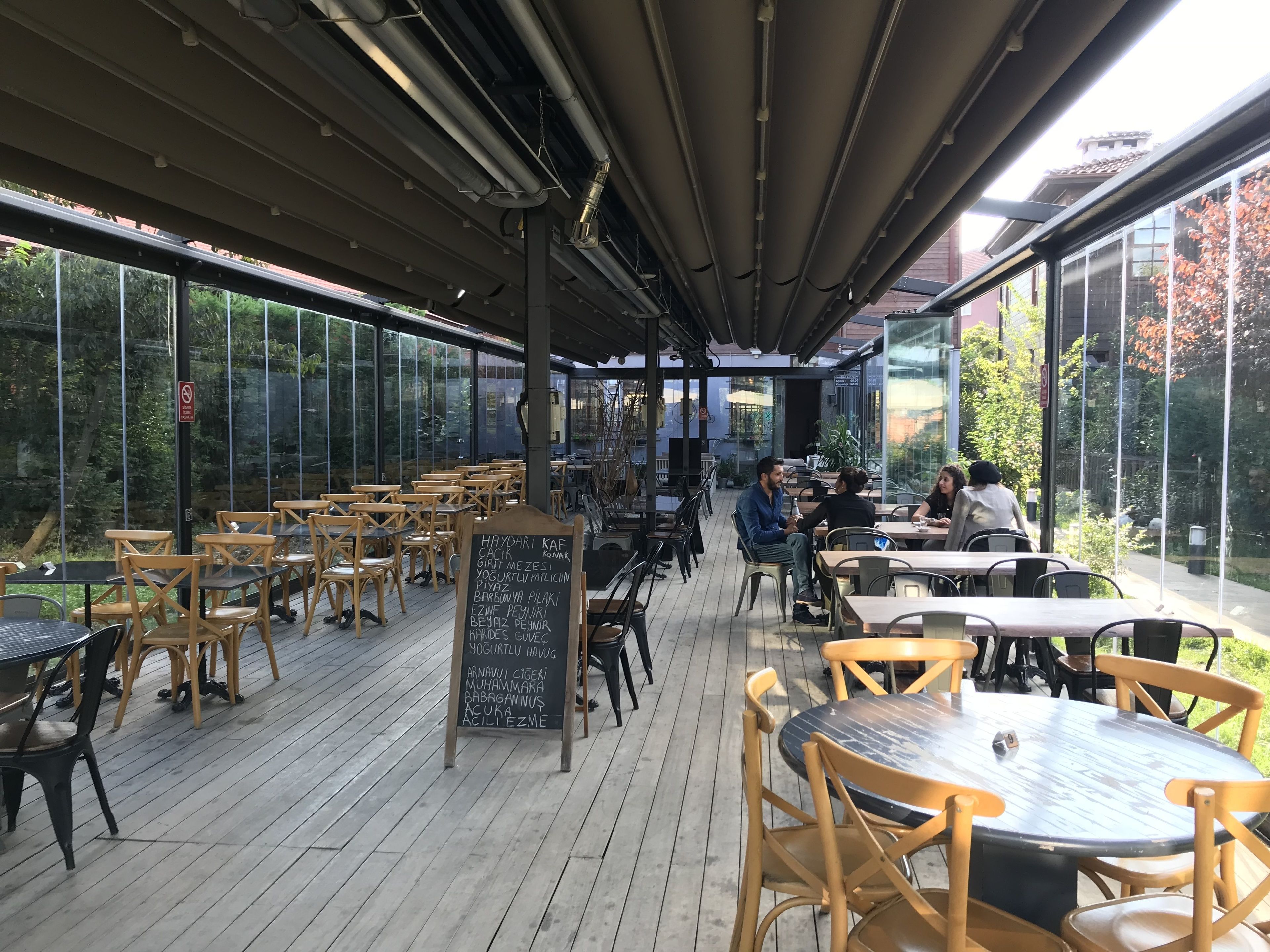 Un café al aire libre con mesas y sillas de madera, un menú en una pizarra y amplios ventanales que ofrecen vistas del entorno verde.