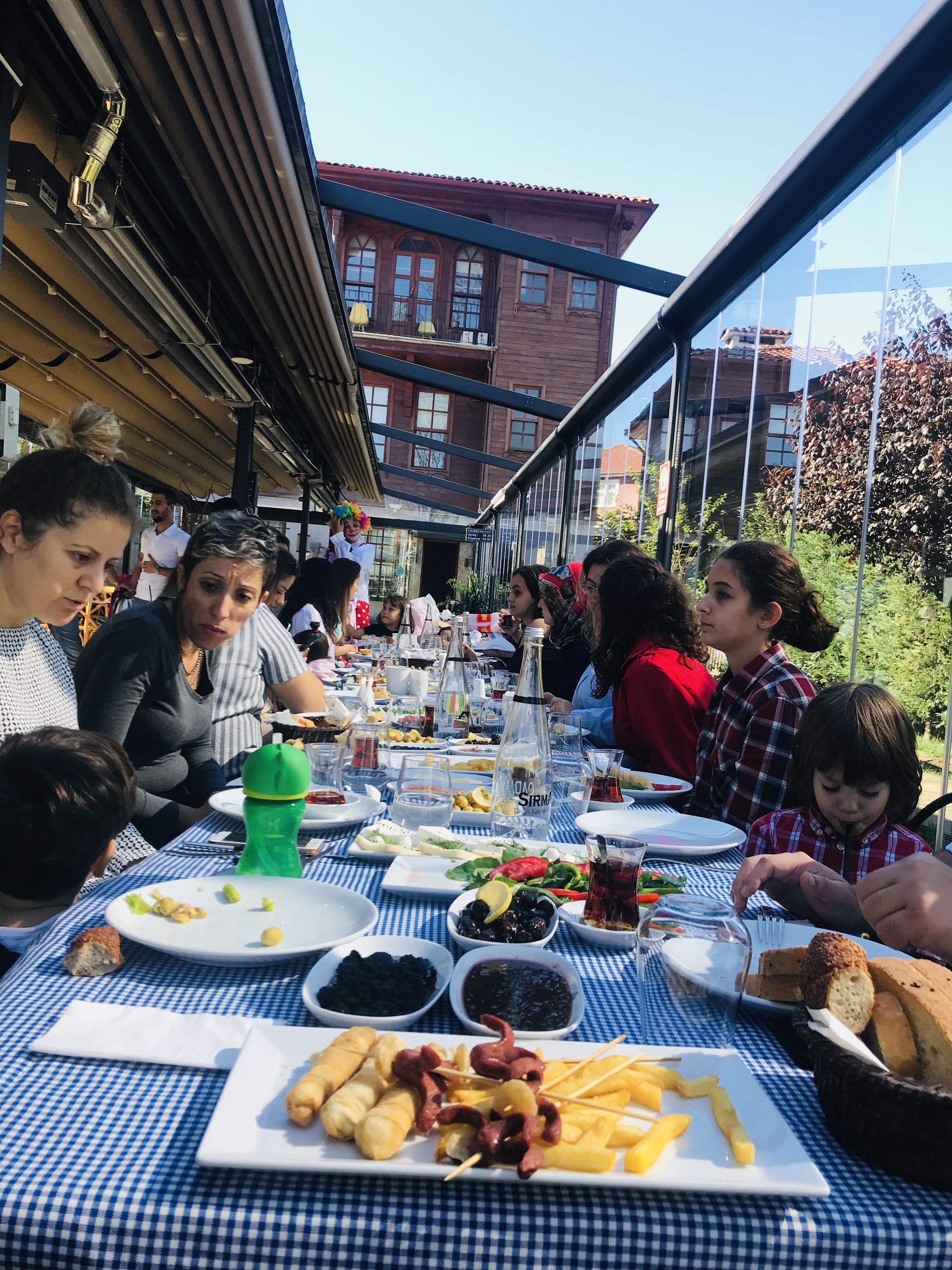 Un grupo de personas cenando en una mesa al aire libre con mantel a cuadros, varios platos y una botella de agua.