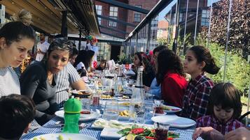 Un grupo de personas cenando en una mesa al aire libre con mantel a cuadros, varios platos y una botella de agua.