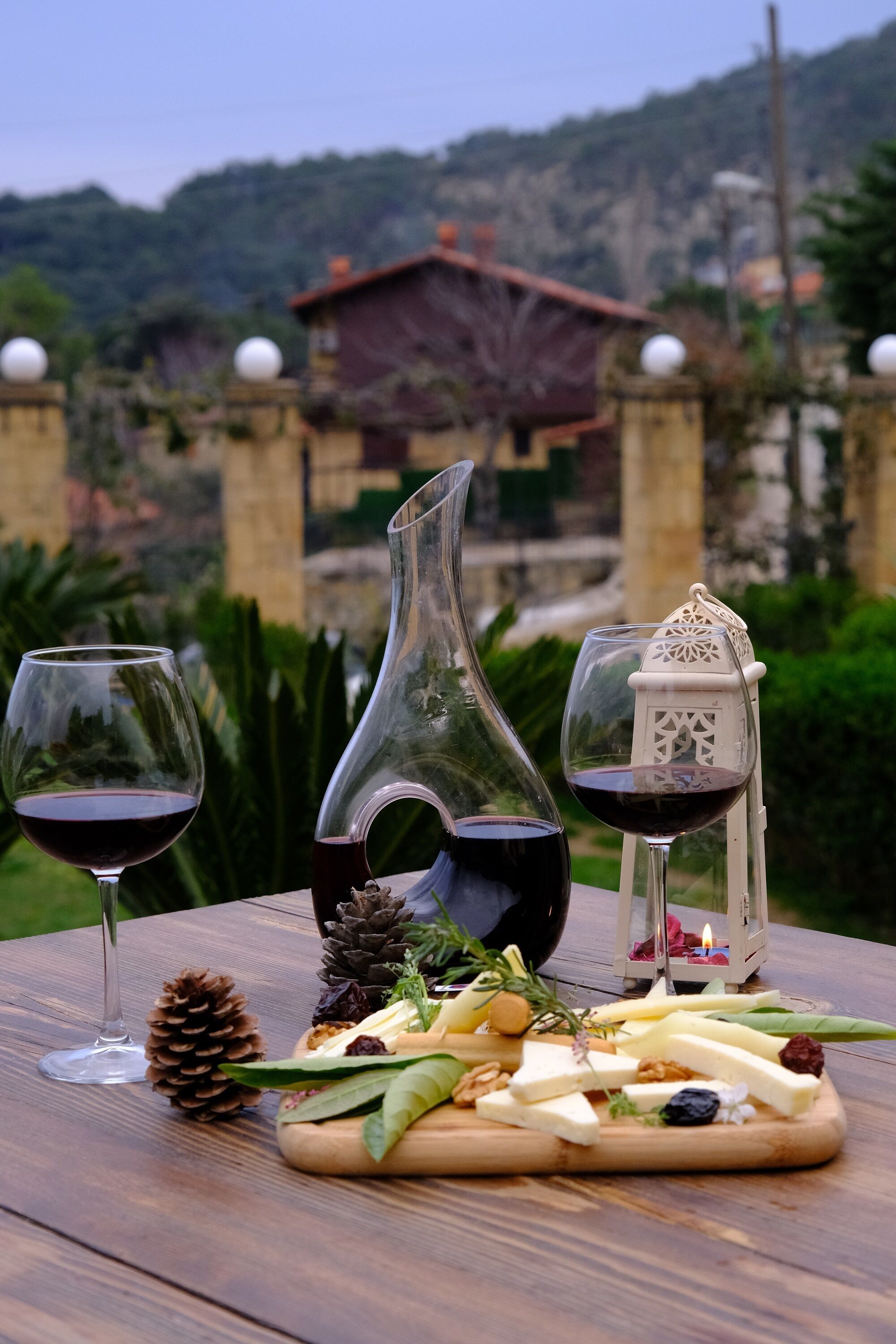 Uma mesa de madeira com uma tábua de queijos, dois copos de vinho tinto, um jarro decorativo e uma vela.