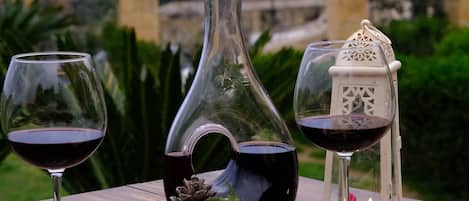 Uma mesa de madeira com uma tábua de queijos, dois copos de vinho tinto, um jarro decorativo e uma vela.